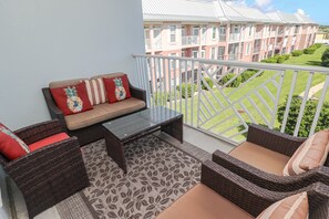 A balcony with wicker furniture, a sofa with floral cushions, and a view of a residential building. - Seaside at Anastasia E302- Renovated Luxury Penthouse Condo (St Augustine)