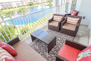 A balcony with wicker furniture, a glass-top table, and a view of a swimming pool and buildings. - Seaside at Anastasia E302- Renovated Luxury Penthouse Condo (St Augustine)