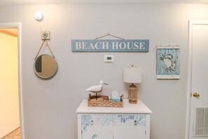 A hallway with a sign reading 'BEACH HOUSE,' a round mirror, a wall clock, a lamp, a decorative bird, and a framed artwork.