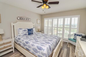 A bedroom with a bed, a nightstand, a lamp, a ceiling fan, a window with blinds, and a sign that reads 'BEACH HOUSE'.