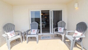 Ein überdachter Sitzbereich mit zwei Adirondack-Stühlen und einem kleinen Tisch, eine Schiebetür zu einer Küche und eine Fußmatte mit dem Aufdruck 'Leave the Sand at the Beach'.