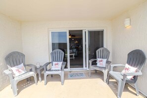 A covered outdoor seating area with two Adirondack chairs and a small table, a sliding glass door leading to a kitchen, and a doormat with 'Leave the Sand at the Beach' text. - Just Steps From the Pool & Beach!! Also has Boat Ramp and Fishing Pier! Trade Winds 51 (St. Augustine)