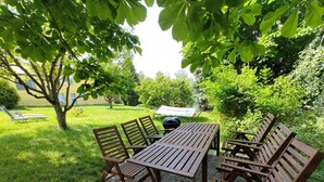 A wooden table and chairs set on a gravel area surrounded by grass and trees, with a hammock and grill in the background. - Haus Nr. 37 (Wunsiedel)
