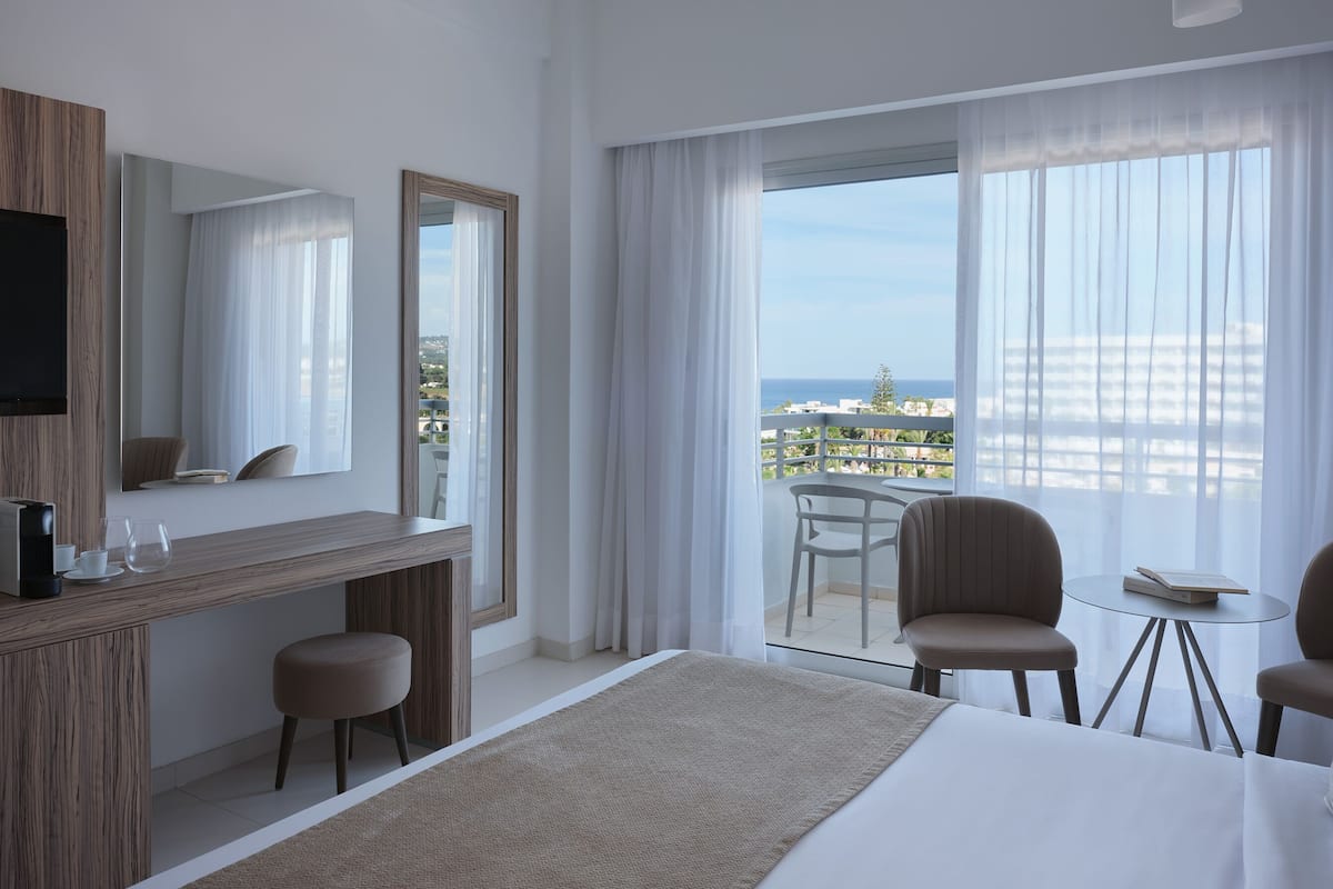 A modern hotel room with a large bed, a desk with a chair, a small table with a chair, and a balcony with a view of the city and ocean.