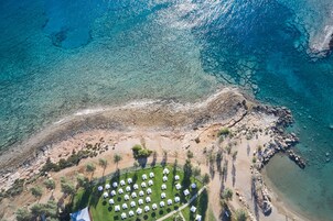 An aerial view of a coastal area with clear blue waters, rocky terrain, and a row of white tents arranged in a semi-circle, suggesting a campsite or outdoor event.