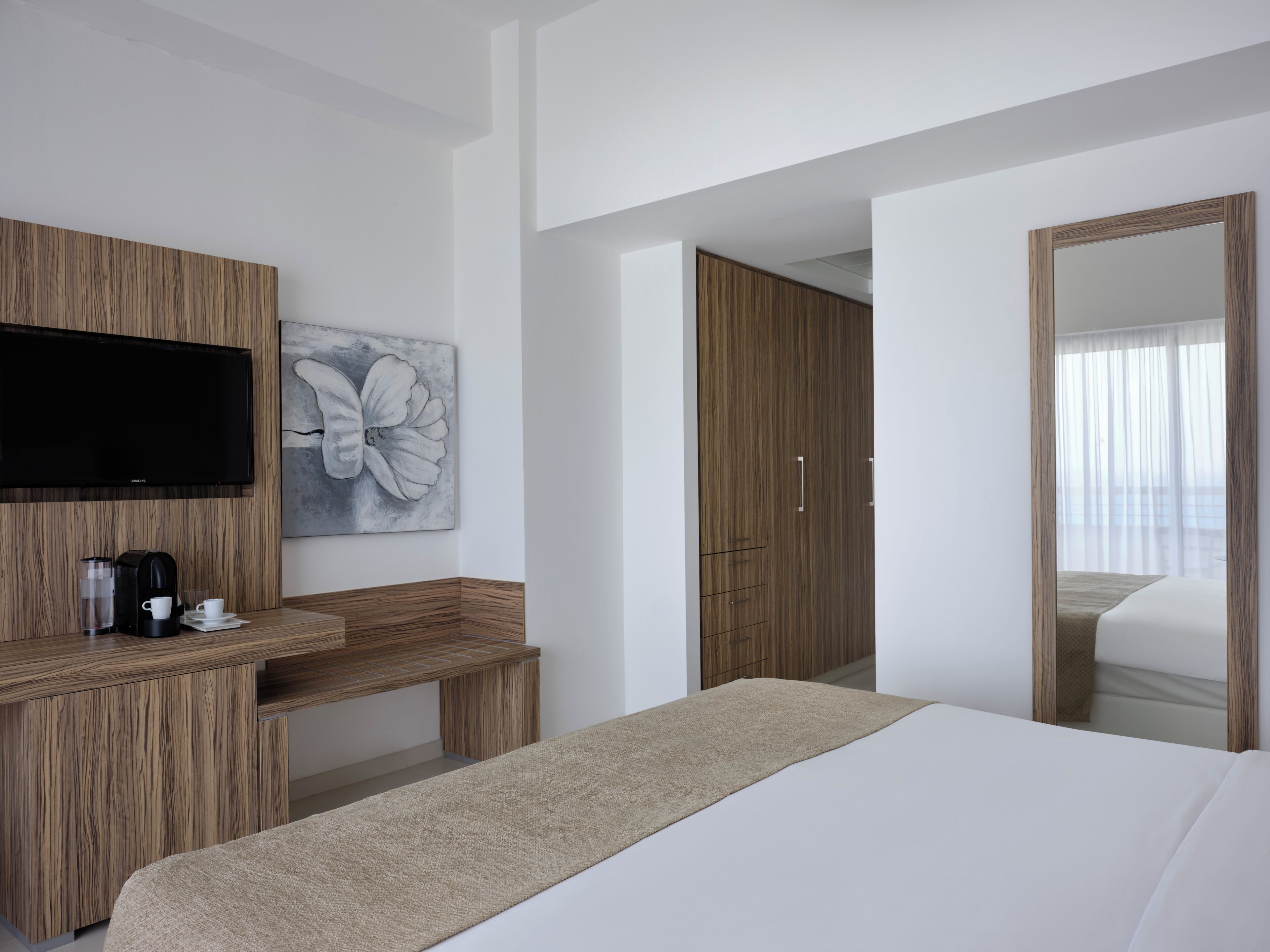Habitación de hotel con cama, televisor montado en un panel de madera, cafetera y un cuadro en la pared.