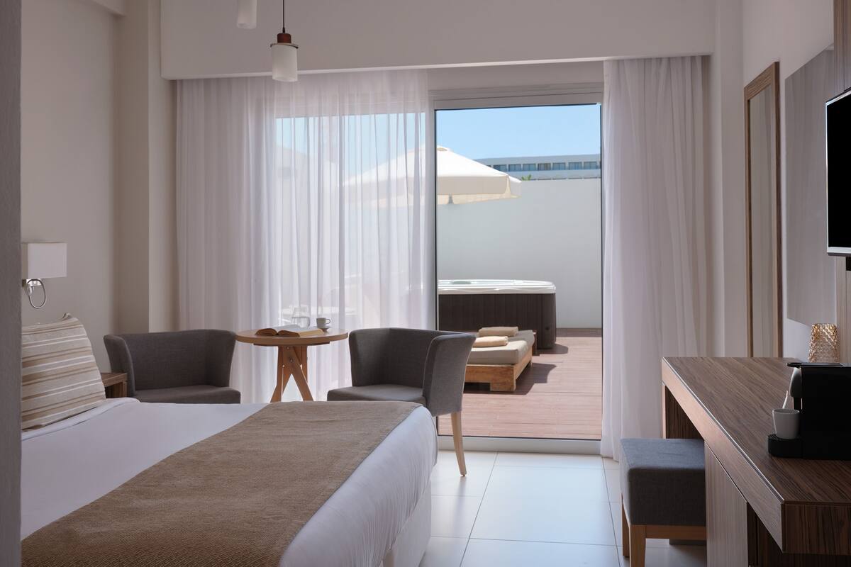 A hotel room with a large bed, a desk with a TV, two chairs, a small table with a cup and a book, and a view of a pool and building through sheer curtains.