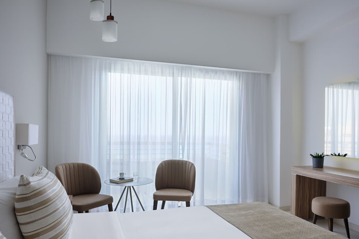 A hotel room with a large bed, two chairs, a small table with a cup and book, a desk with a plant, and sheer curtains.