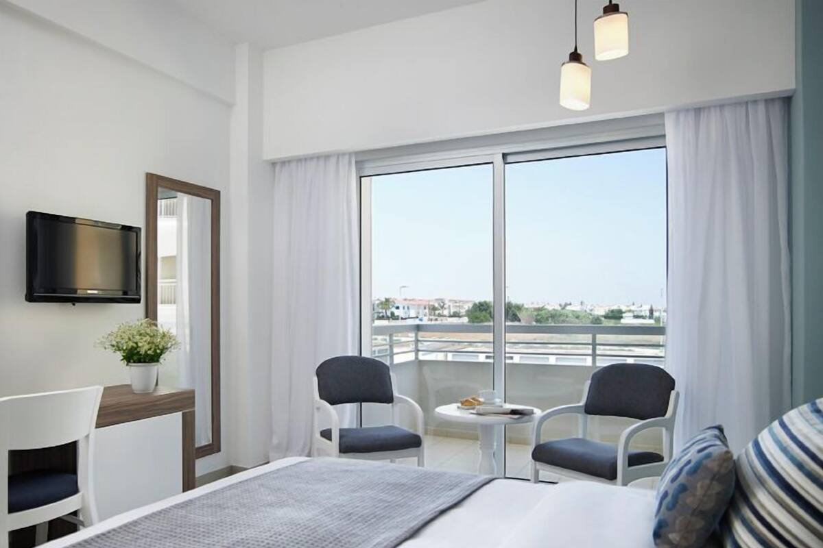 A hotel room with a bed, two chairs, a small table, a TV, and a large window with curtains.