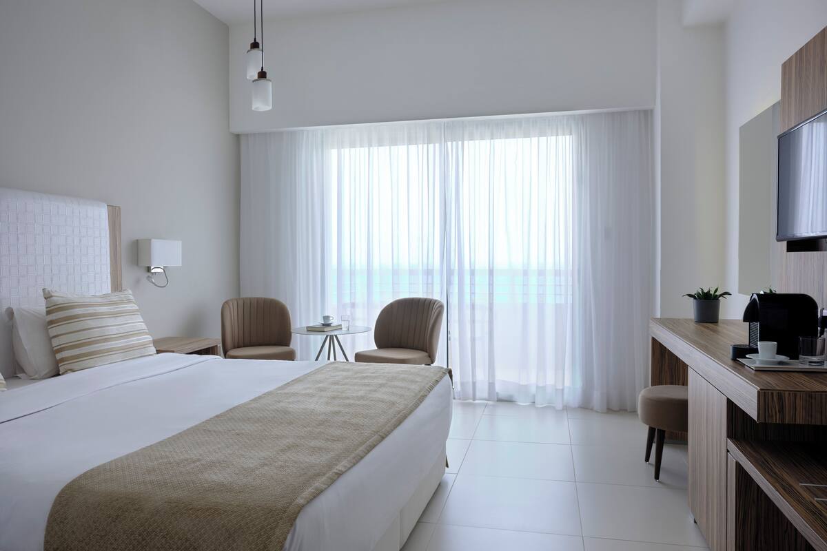 A hotel room with a large bed, two chairs, a small table with a cup and book, a desk with a coffee maker, and a TV.