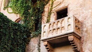 Un edificio histórico con un balcón que presenta ornamentación de piedra labrada y fachada de ladrillo, parcialmente cubierta de hiedra verde.