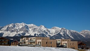Un hotel moderno con varios pisos y balcones, con vistas a montañas nevadas bajo un cielo azul despejado.