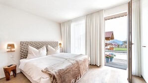 Un dormitorio con una cama grande, una mesita de noche de madera, un cabecero con diseño estampado y vistas a un edificio y montañas a través de la ventana.