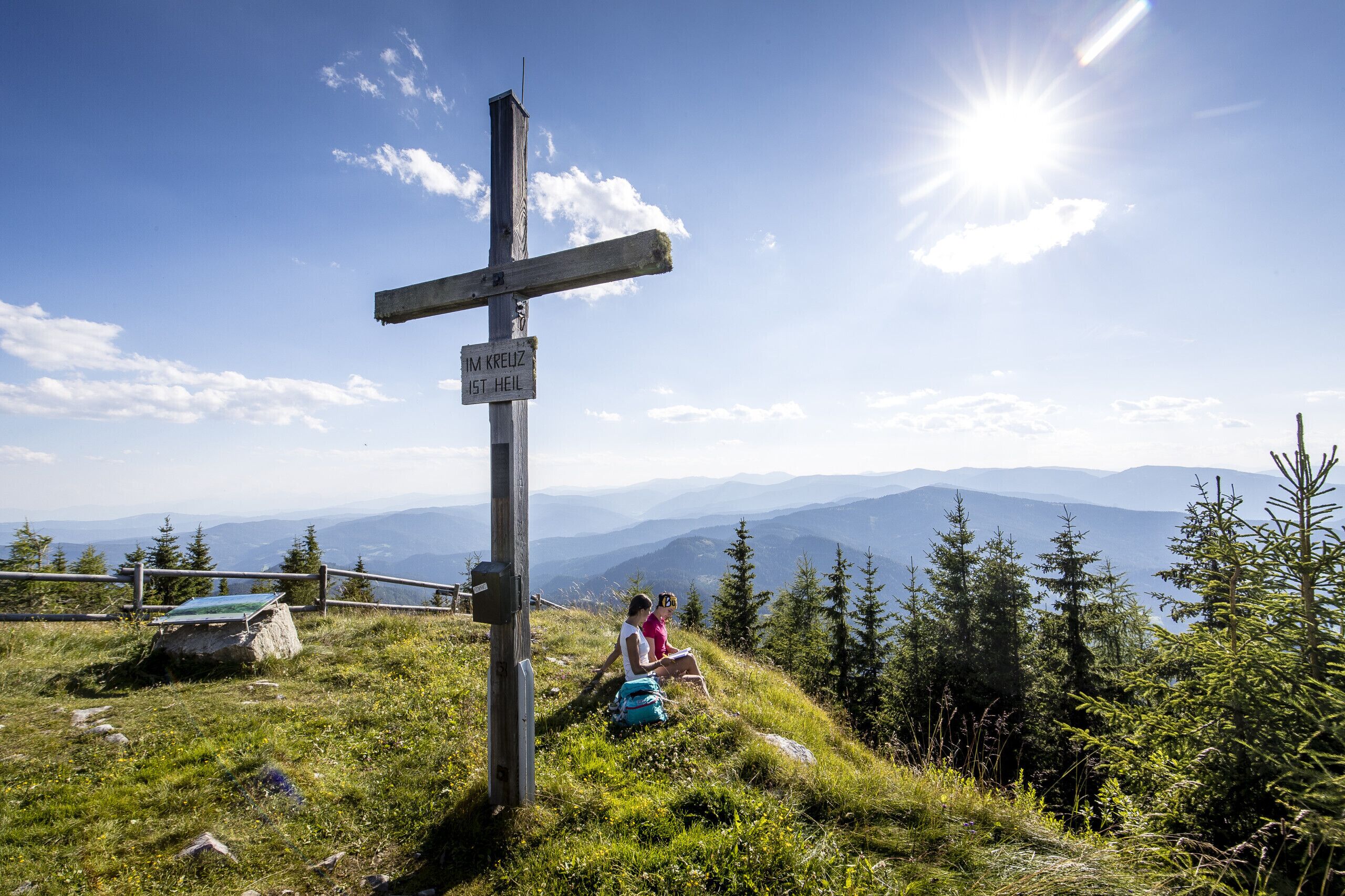 A wooden cross with a sign, surrounded by a grassy area with trees and mountains in the background.