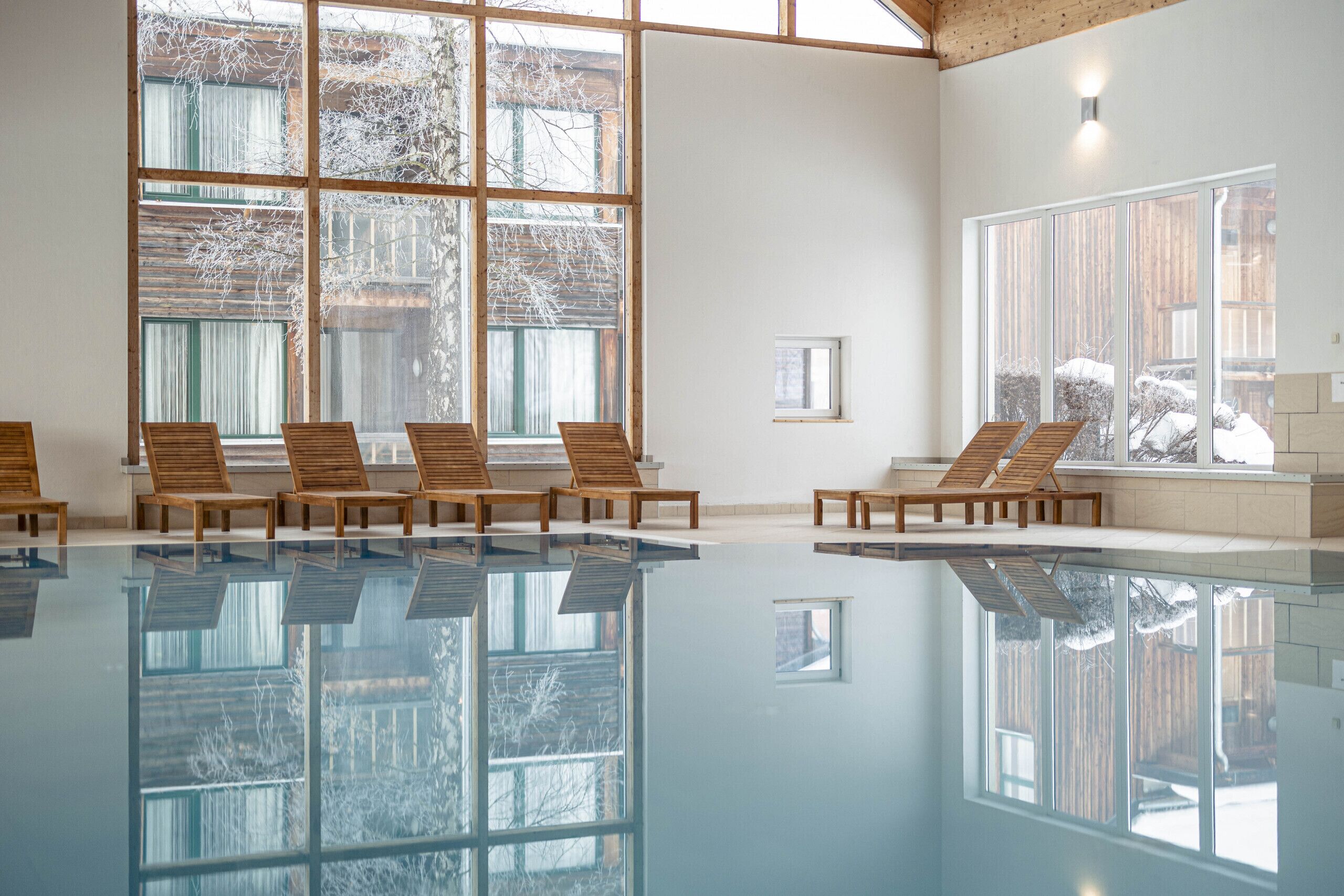 A large indoor swimming pool with clear glass walls, wooden lounge chairs, and large windows offering views of a snowy landscape.