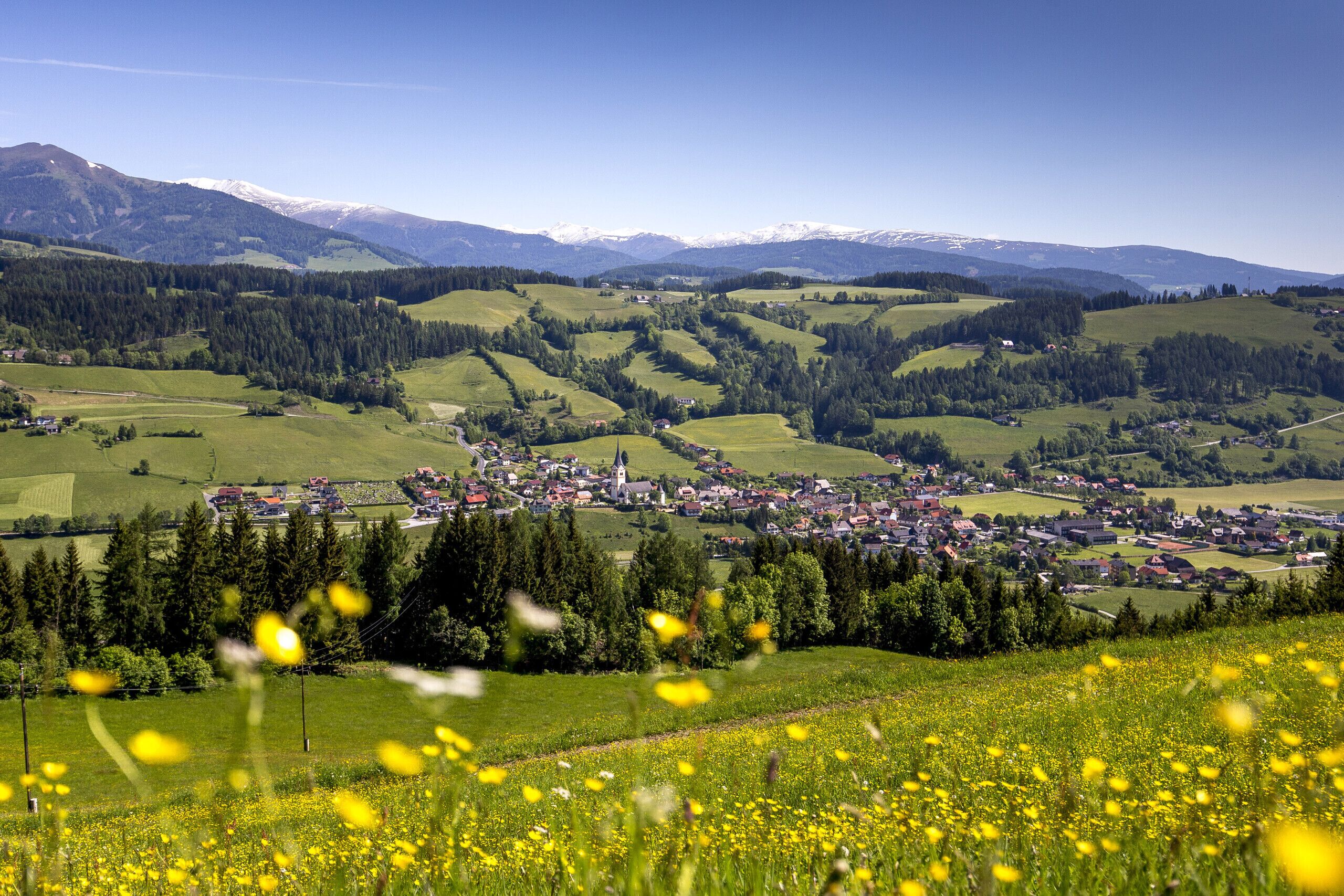 A rural town nestled in a valley with rolling hills and mountains in the background.