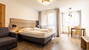 A modern hotel room with a wooden headboard, a bed with white linens, a grey sofa, a wooden dining table with chairs, and a desk with a lamp.