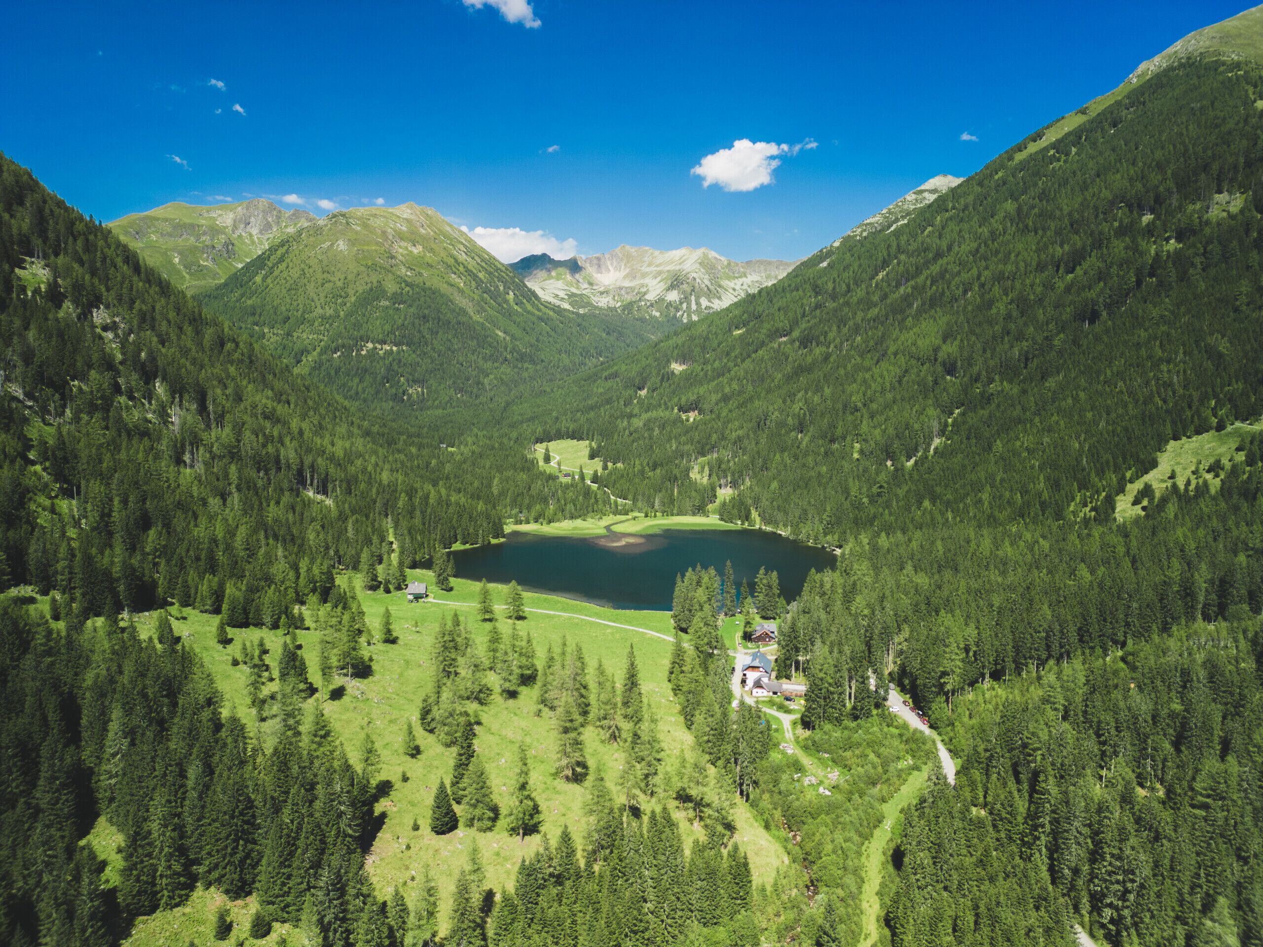 Un paysage montagneux avec un lac, des zones boisées et un petit bâtiment doté d’un toit rouge.