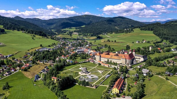 Luftaufnahme einer Stadt mit einem markanten, schlossartigen Gebäude, umgeben von grünen Feldern und Bergen.