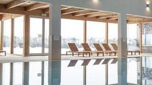Ein moderner Poolbereich im Innenbereich mit Holzbalken, großen Fenstern und Liegestühlen.