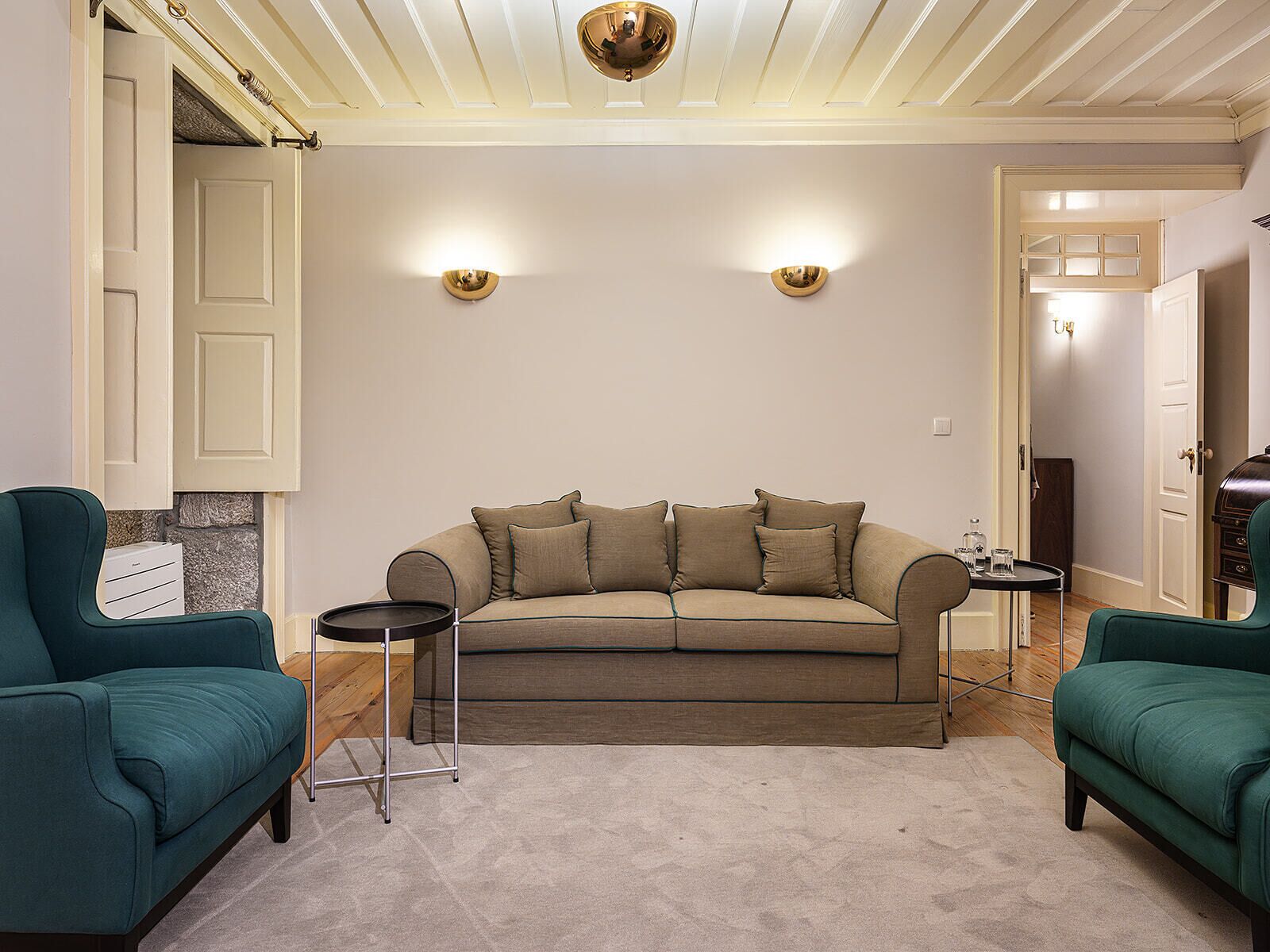 Een woonkamer met een beige bank, twee groene fauteuils, een klein bijzettafeltje en een decoratieve spiegel.