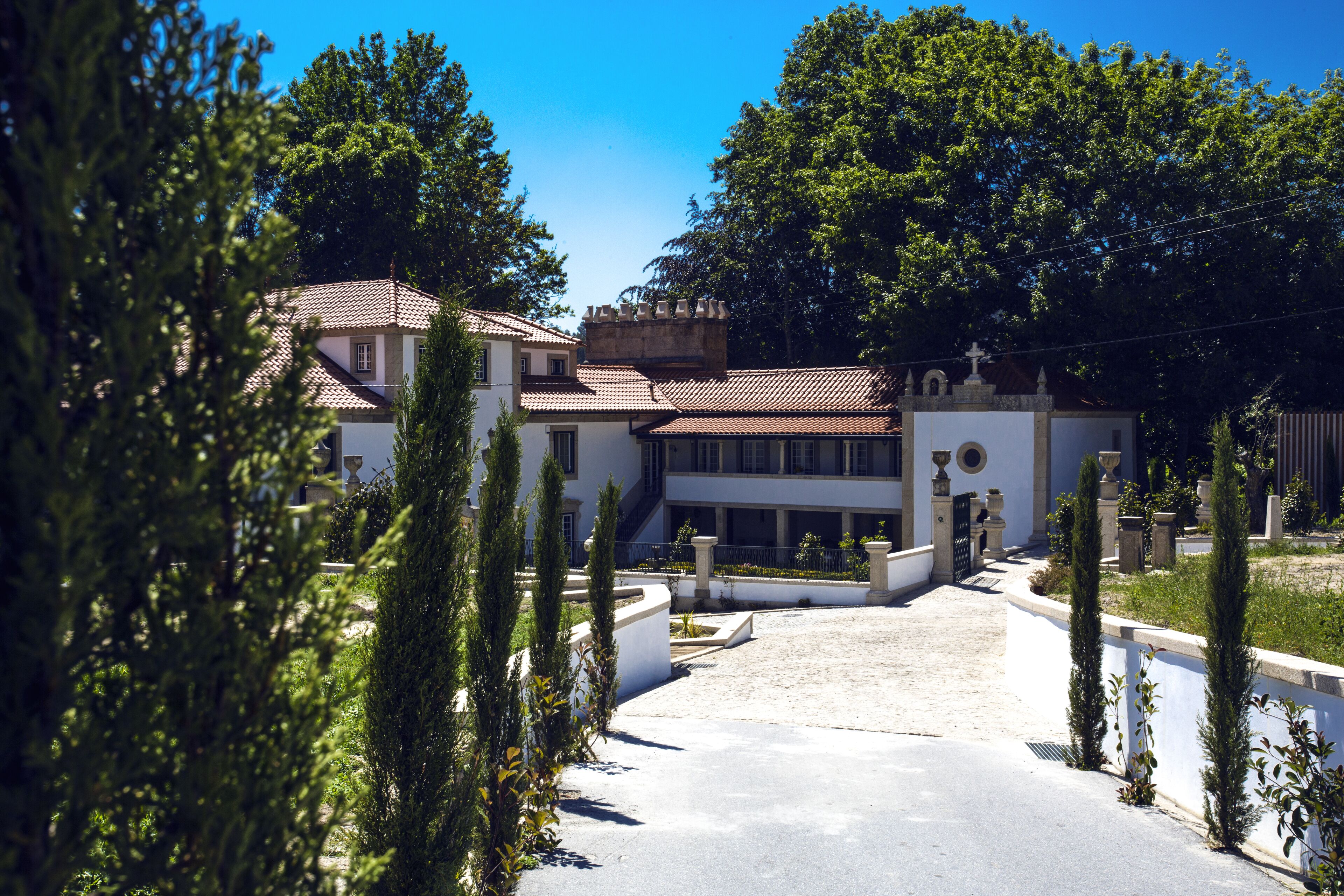 A large, white building with a tiled roof, surrounded by greenery and a paved driveway.