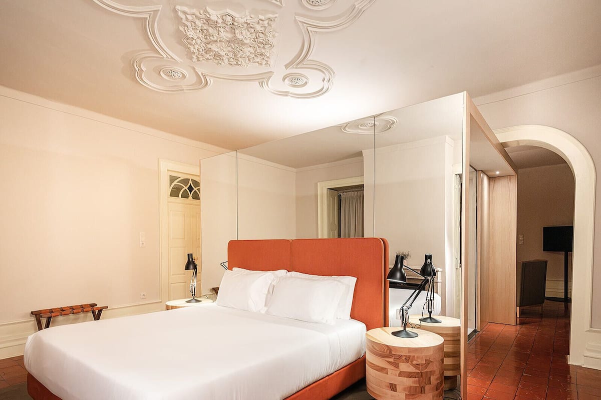 A spacious bedroom with a large bed, a wooden nightstand, a mirror, and a door leading to another room.