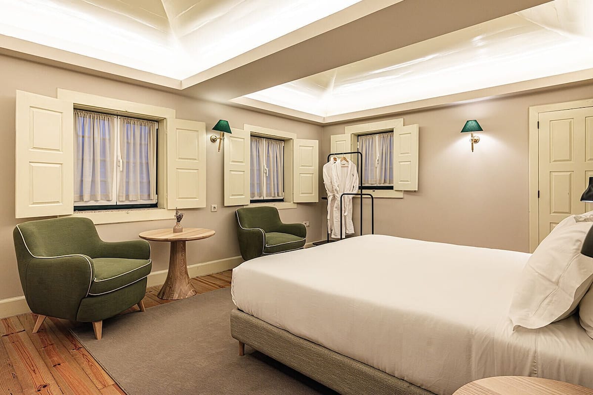 A hotel room with a large bed, two green armchairs, a small round table, and a robe rack.
