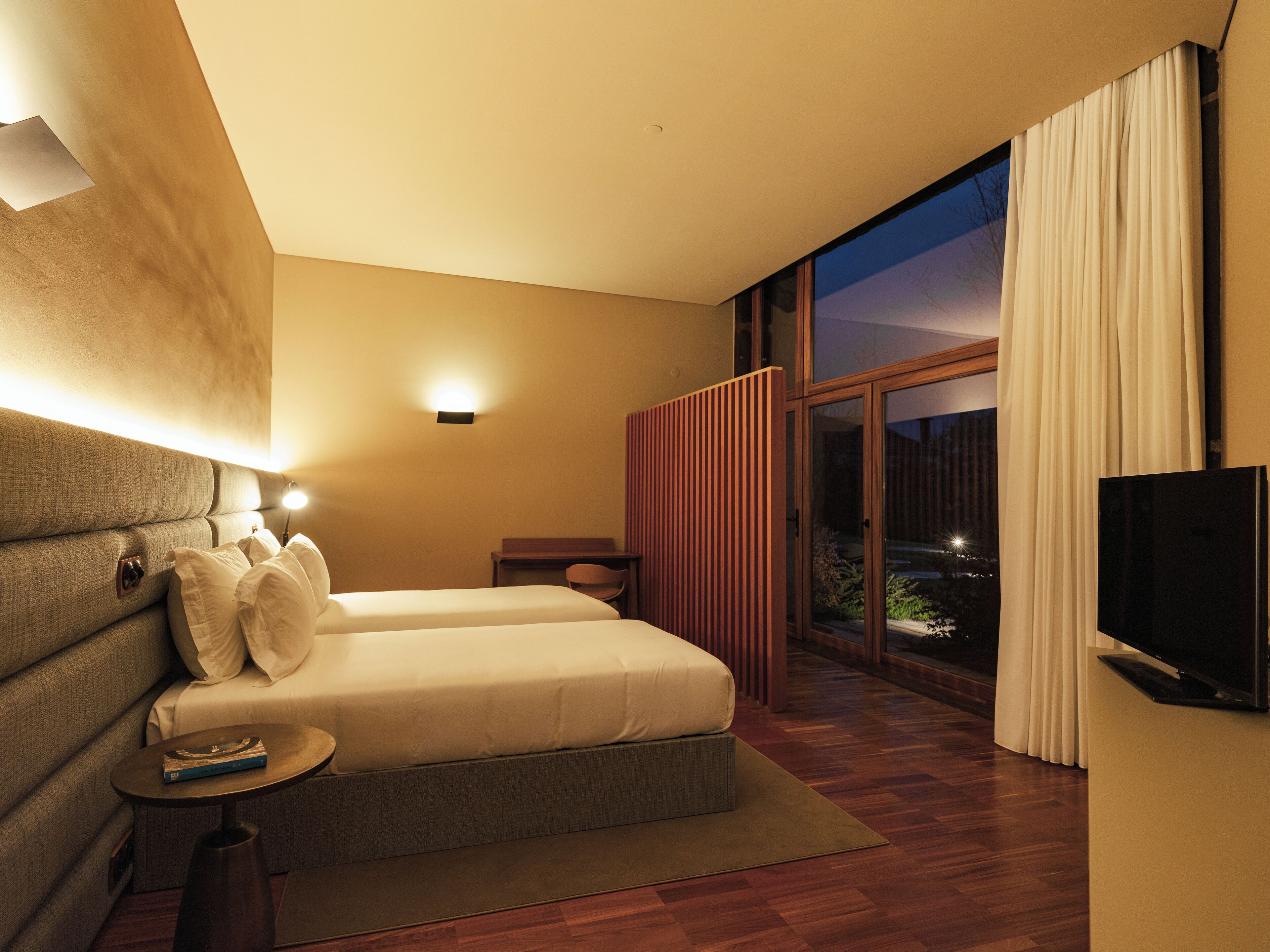 A modern hotel room with a large bed, a small table, a TV, and a window with curtains.