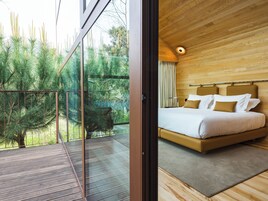 Un dormitorio con techo de madera, cama con ropa de cama blanca y marrón, y vista a árboles a través de un ventanal.
