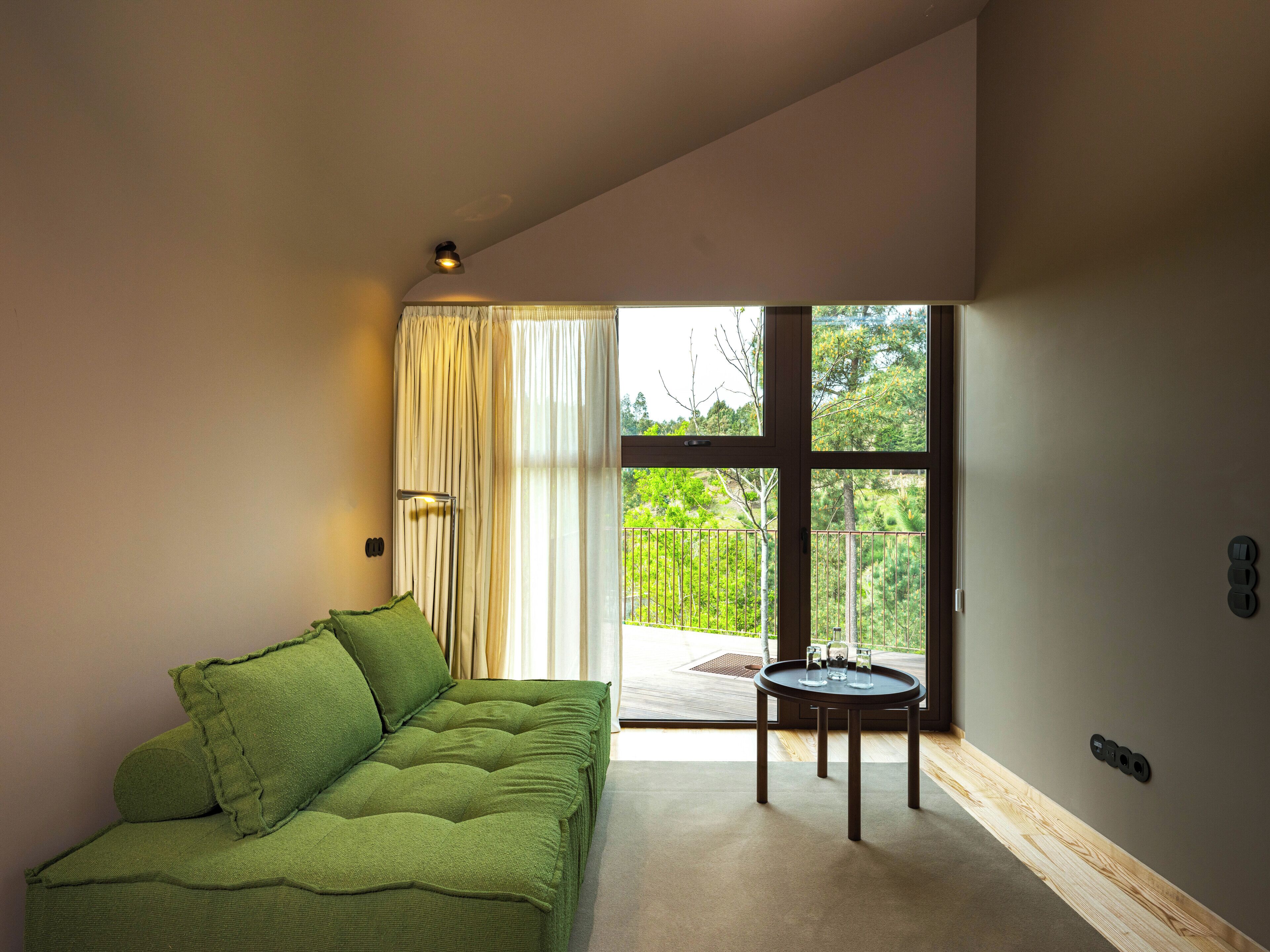 A room with a green sofa, a small round table, and a large window with a view of greenery outside.
