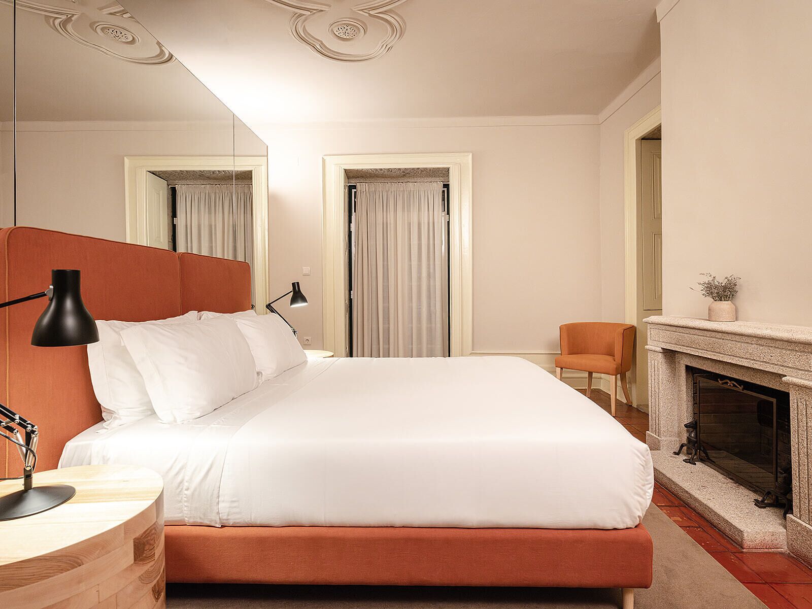 A modern hotel room with a large bed, a desk with a lamp, a chair, and a fireplace.