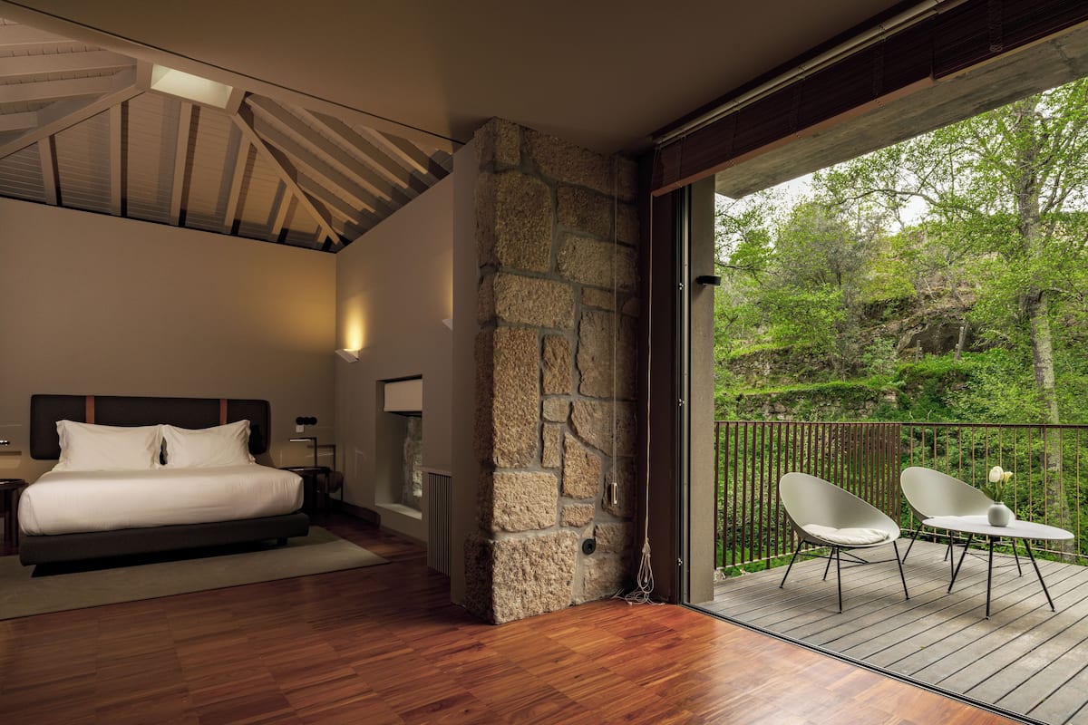 A modern bedroom with a large bed, a stone accent wall, and a balcony with two white chairs.