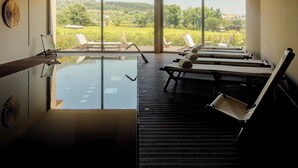 Un área de piscina interior moderna con paredes de vidrio transparente, sillones de descanso y vistas a un paisaje exuberante.