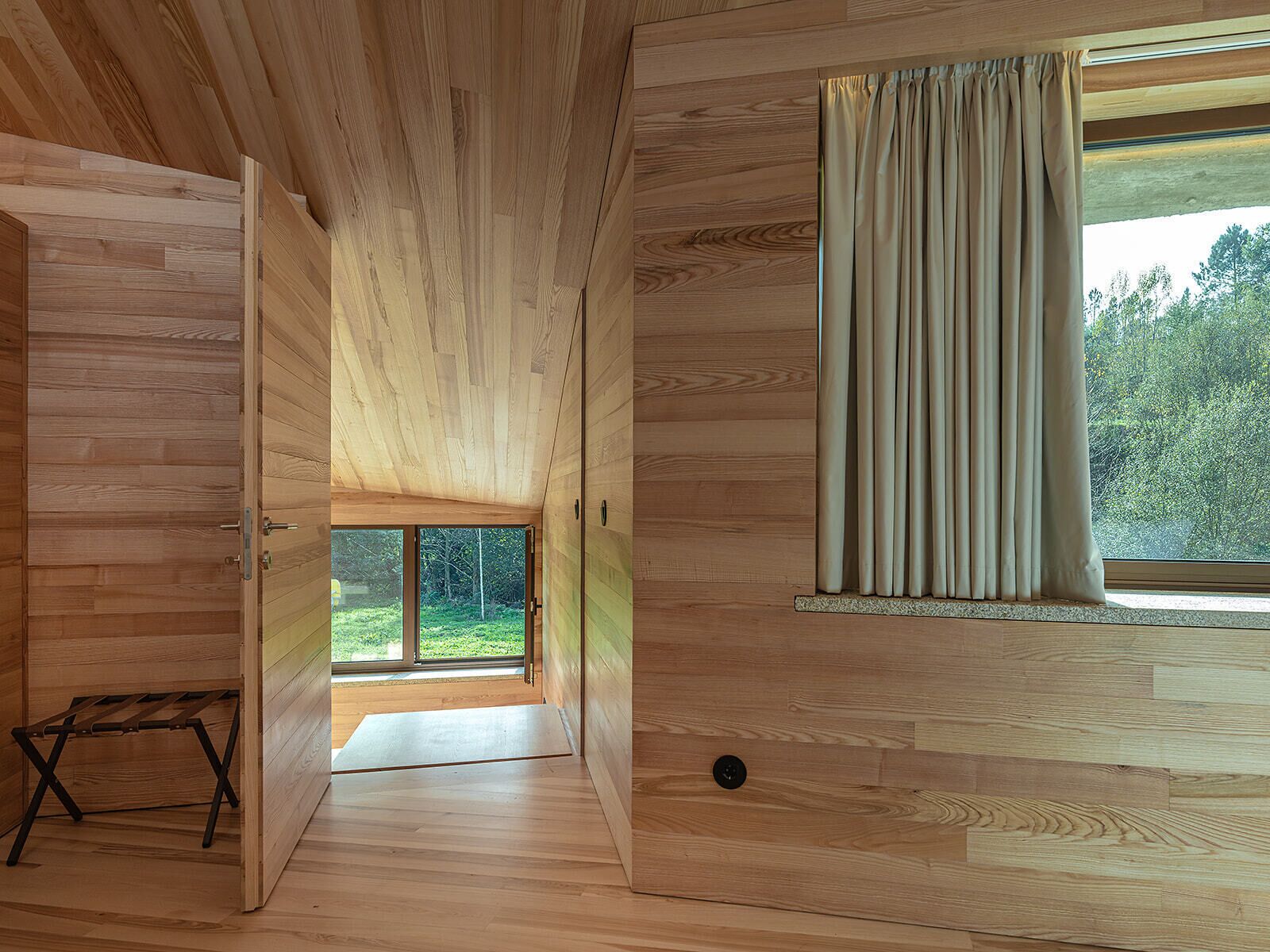 A wooden interior with a large window, a door leading outside, and a small wooden bench.