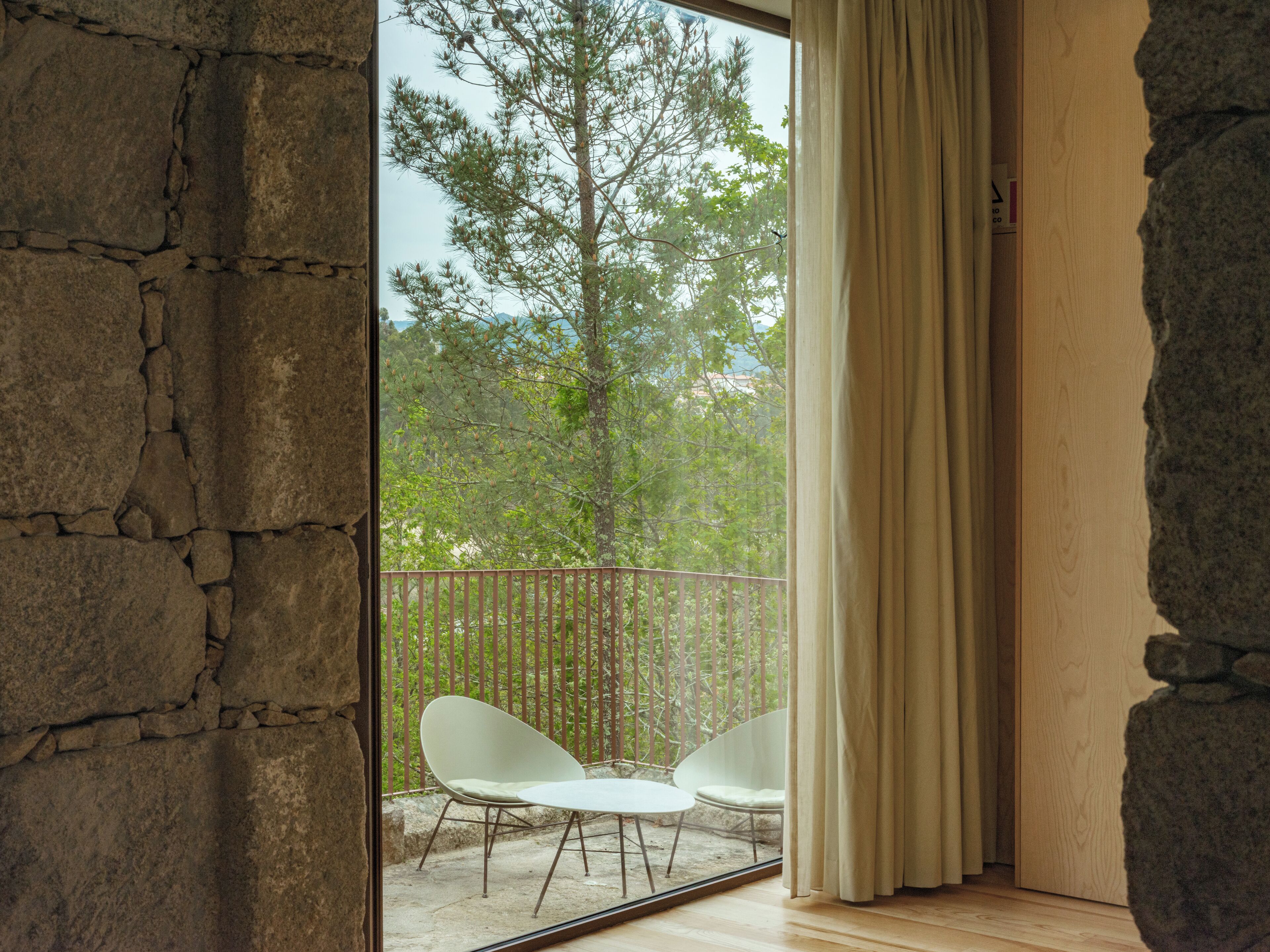 A room with a stone wall, a balcony with two white chairs, and a view of trees and mountains through a window.