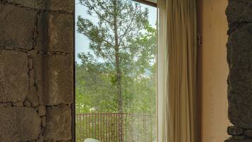 A room with a stone wall, a balcony with two white chairs, and a view of trees and mountains through a window.
