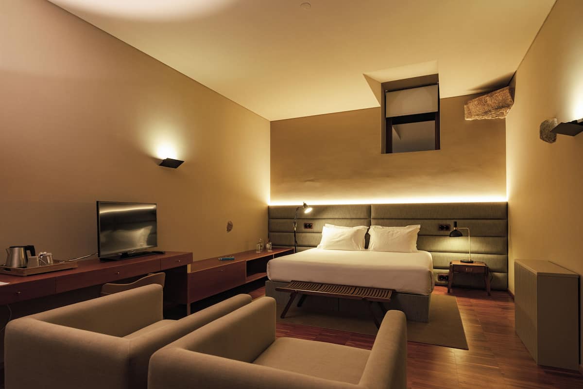 A hotel room with a bed, a desk, a TV, and two armchairs.