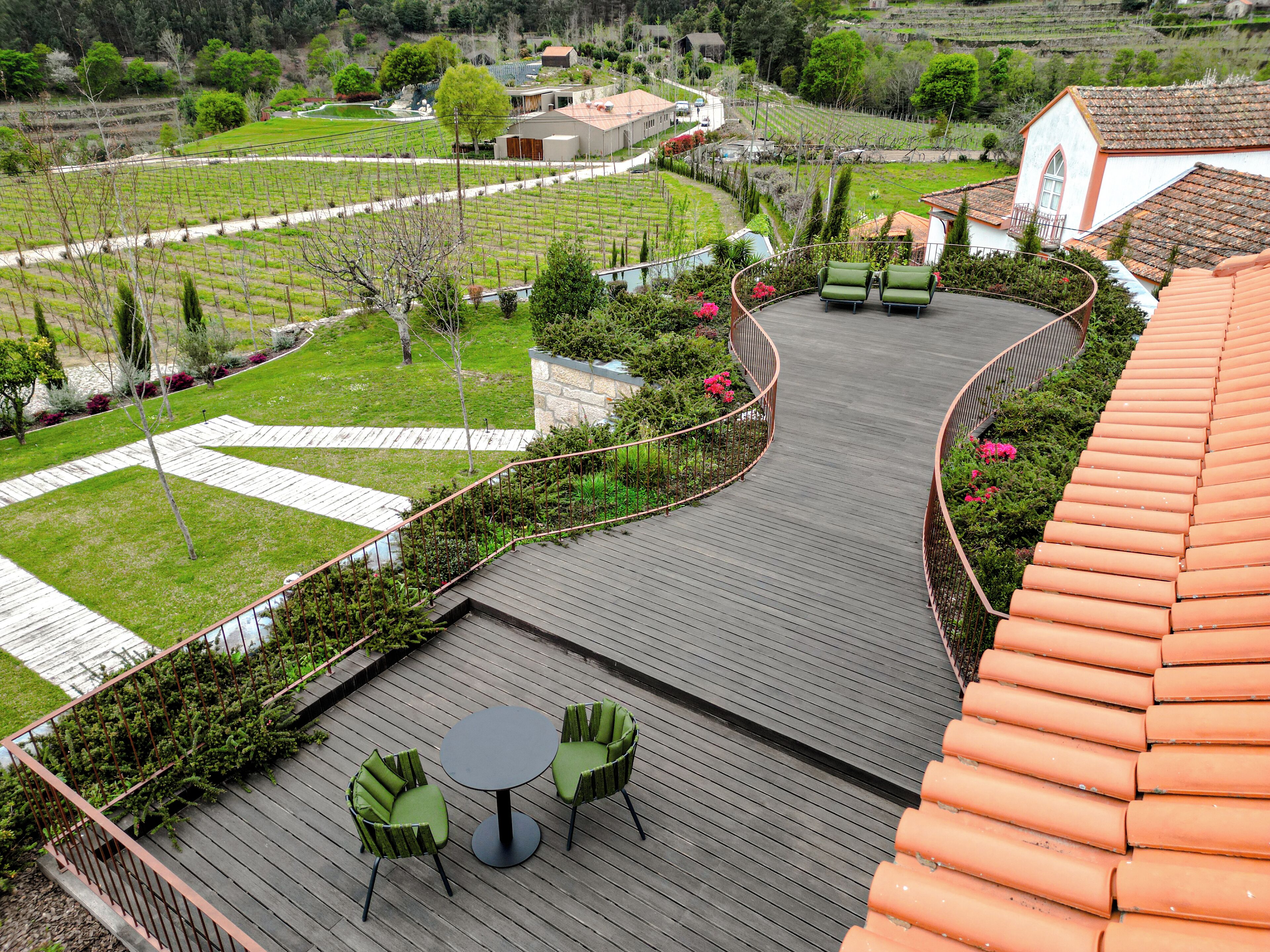 Una terraza de madera con dos sillas verdes y una mesa, rodeada por un jardín con una barandilla curva.