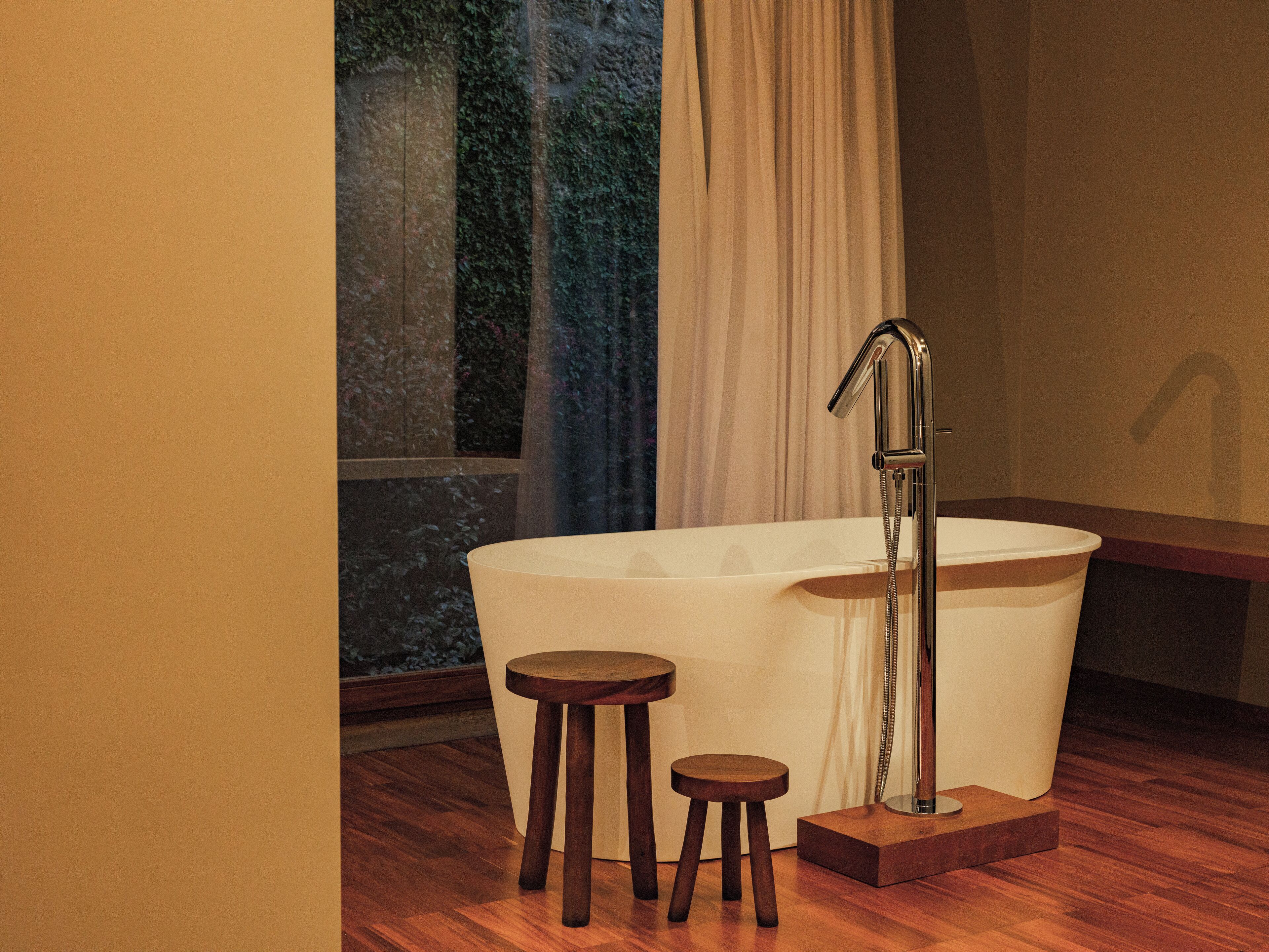 A modern bathroom with a freestanding bathtub, two wooden stools, and a shower head.