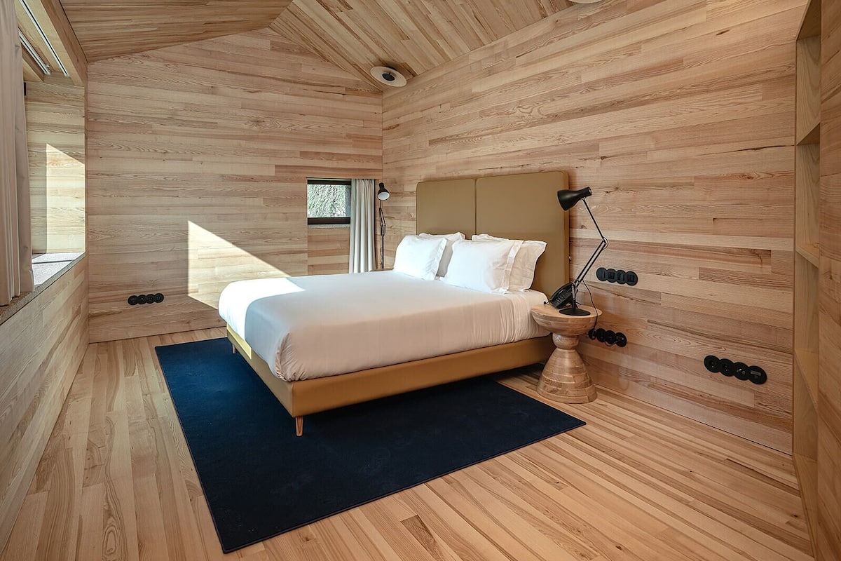 A modern bedroom with a wooden ceiling, a bed with white linens, a bedside lamp, and a small window.