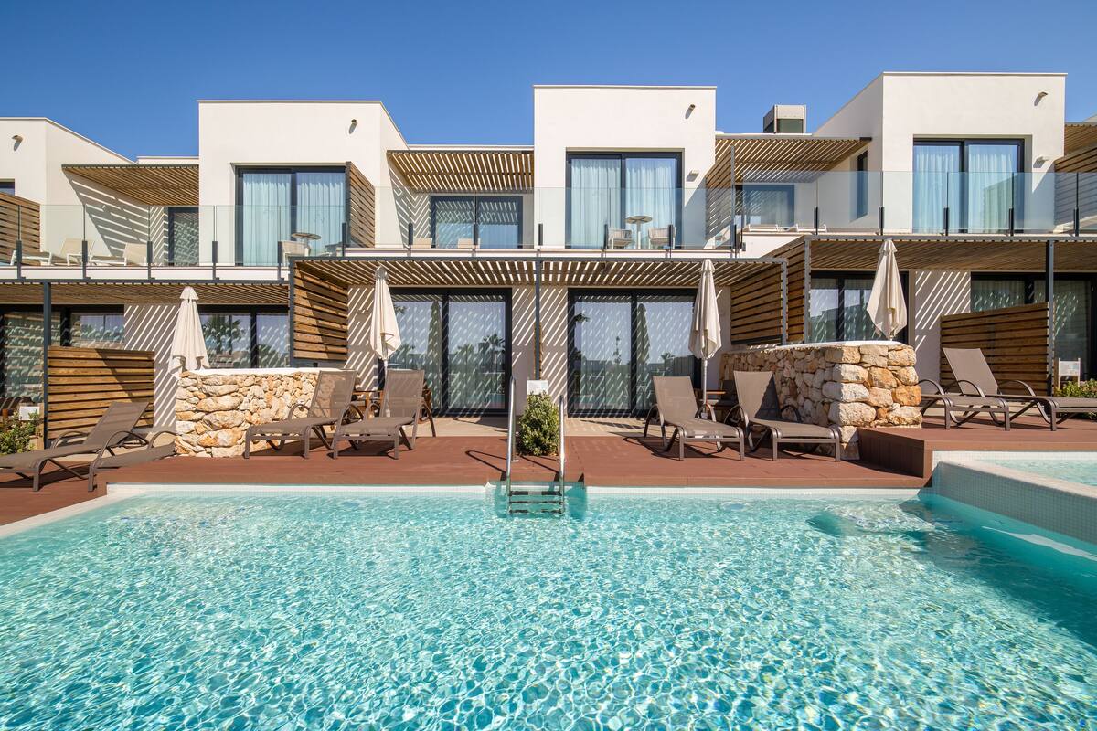 Un hotel moderno con zona de piscina, que incluye sillas de descanso y sombrillas.