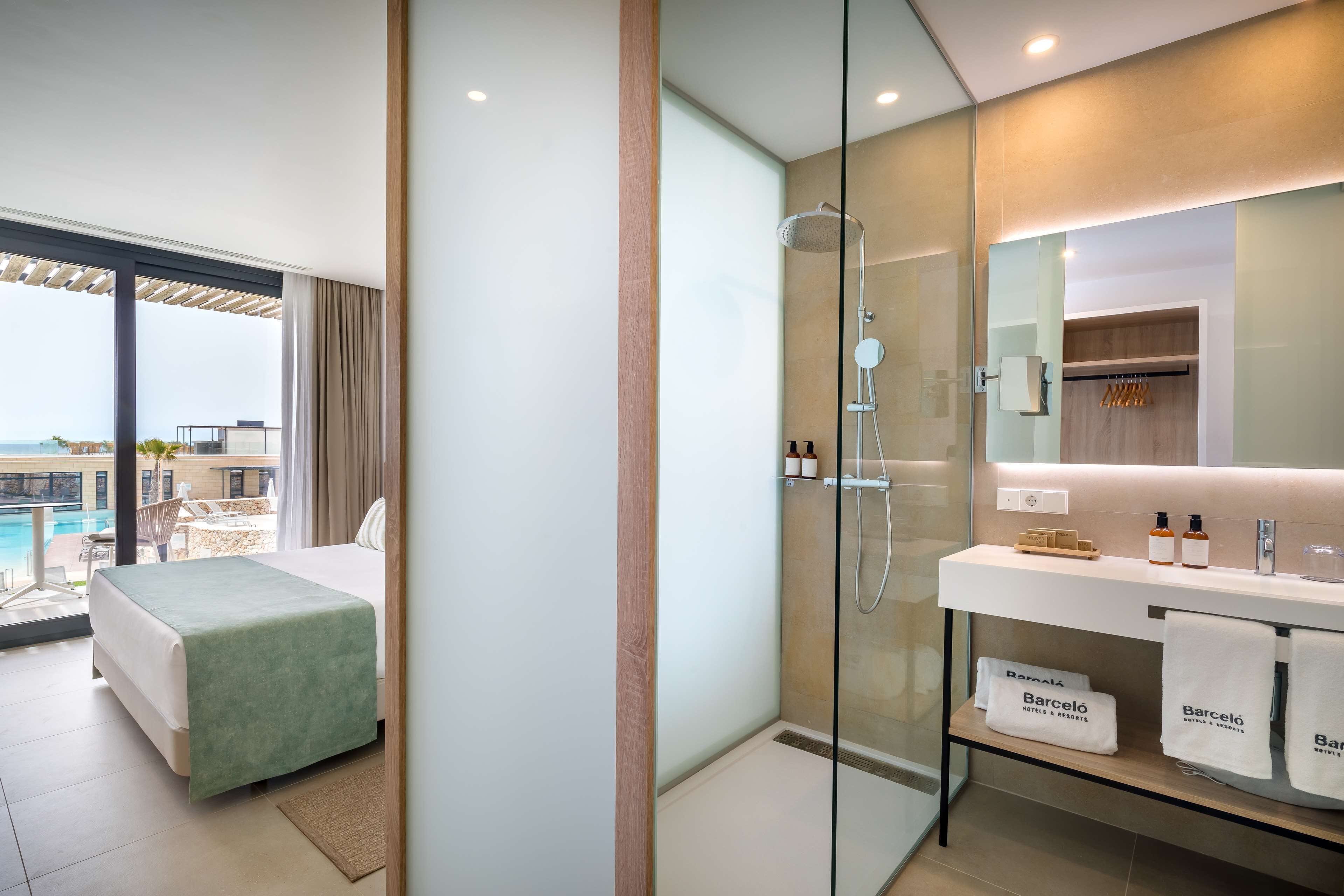 Une chambre d’hôtel moderne équipée d’un lit, d’une douche en verre, d’une coiffeuse avec des articles de toilette et offrant une vue sur la piscine et l’immeuble.