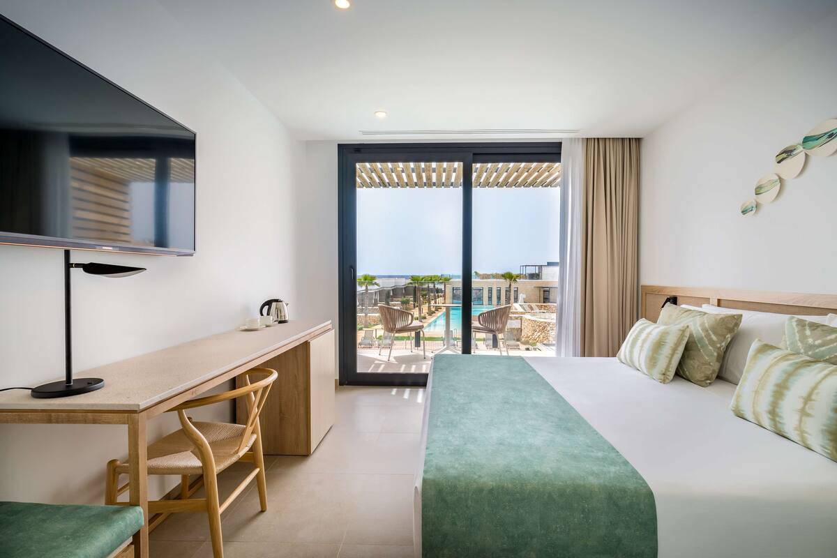Habitación de hotel con cama, escritorio, silla, televisor y vista a una piscina y zona exterior.