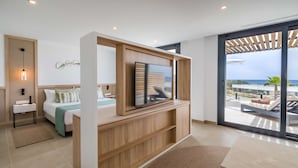 Una habitación de hotel moderna con una cama, un televisor montado en una consola de madera, y un amplio ventanal corredero que da a un balcón con vistas al mar.