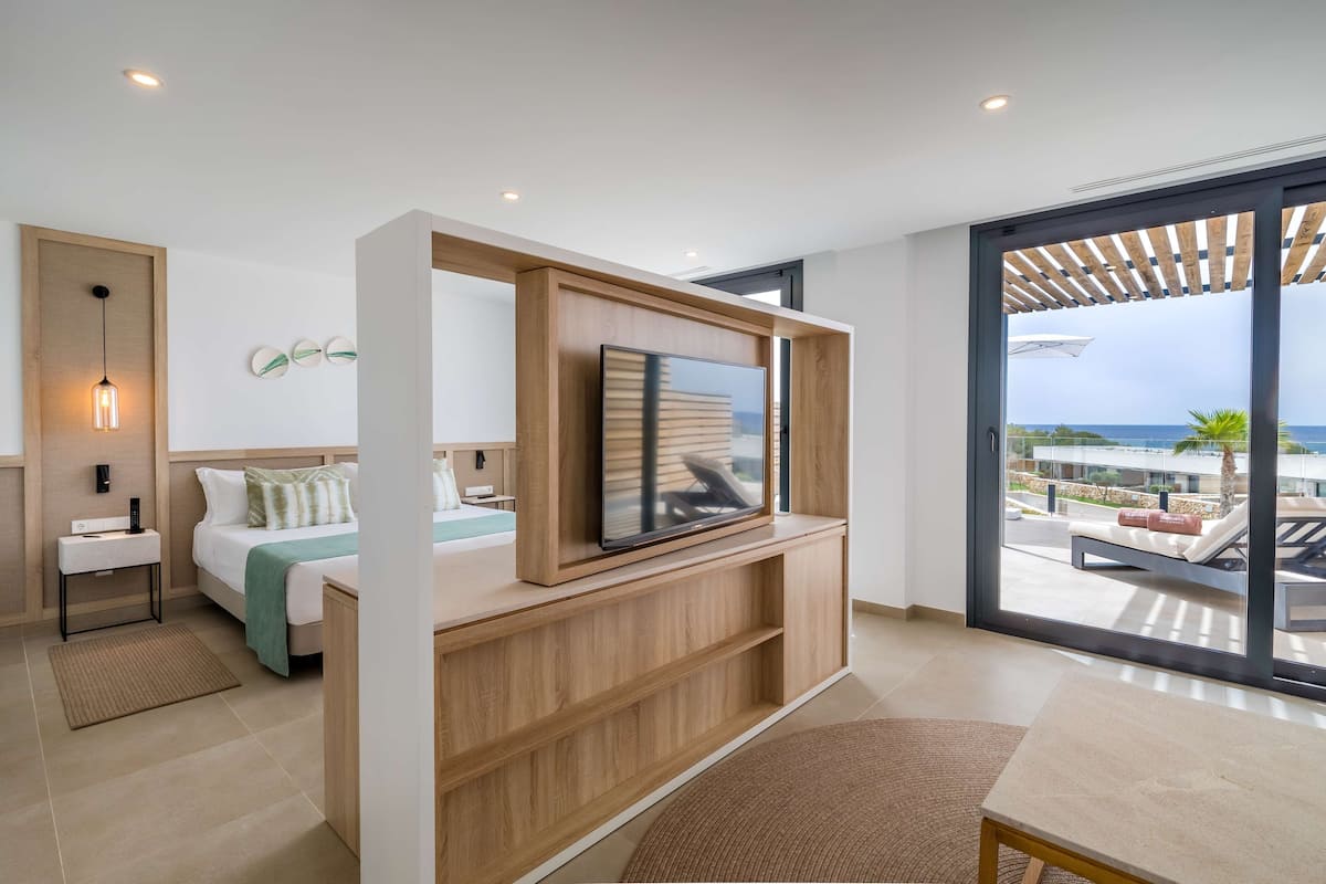 Una habitación de hotel moderna con una cama, un televisor montado en una consola de madera, y un amplio ventanal corredero que da a un balcón con vistas al mar.