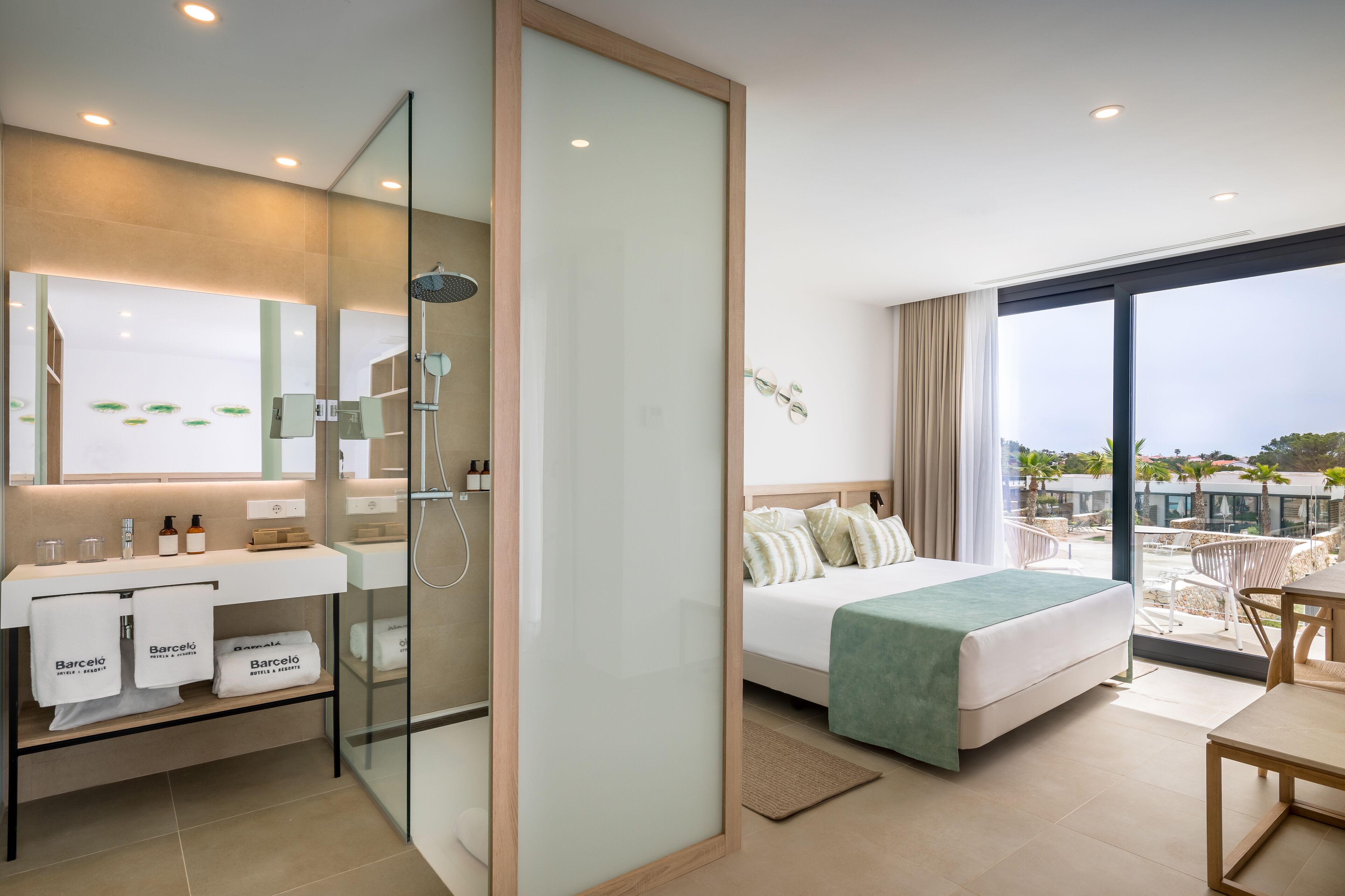 Ein modernes Hotelzimmer mit Dusche in Glasabtrennung, einem großen Bett mit grüner Tagesdecke, einem Balkon mit Aussicht und einer Minibar mit Pflegeprodukten.