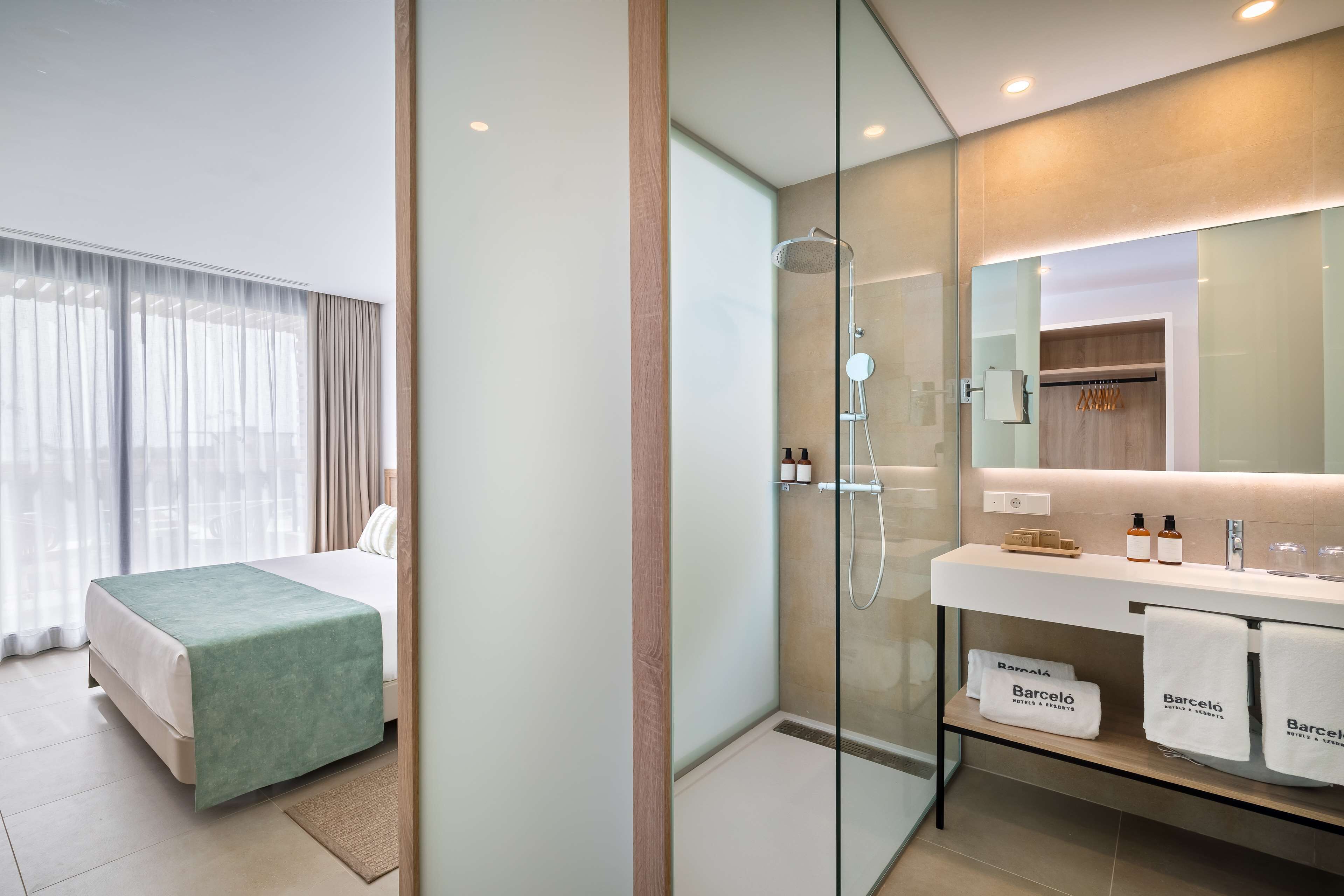 Una habitación de hotel moderna con cama, ducha con mampara de vidrio, tocador con artículos de aseo y vistas a la ciudad.