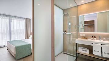 Una habitación de hotel moderna con cama, ducha con mampara de vidrio, tocador con artículos de aseo y vistas a la ciudad.