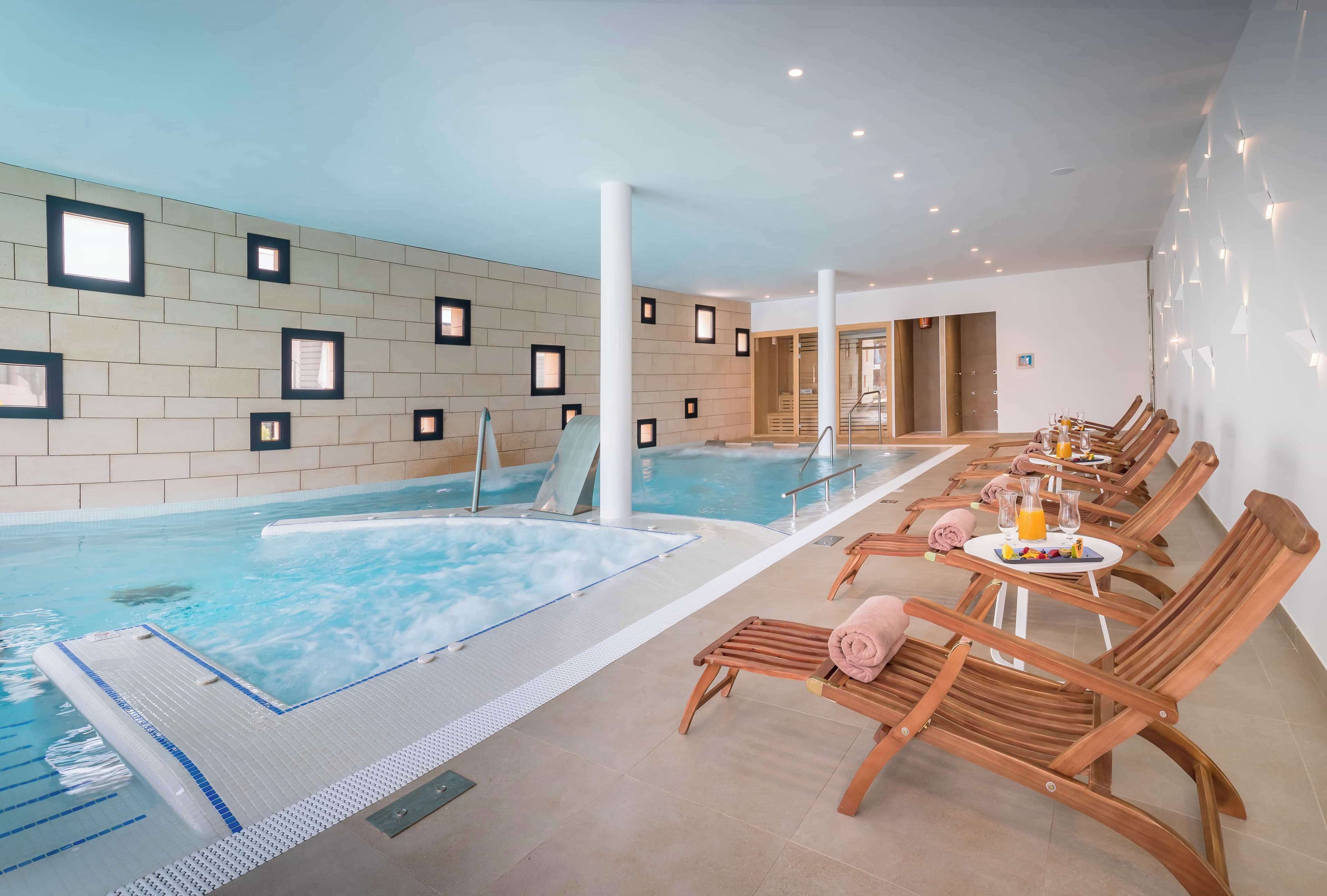 Un espace spa comprenant une grande piscine intérieure, des chaises longues en bois et une table avec des boissons et des serviettes.